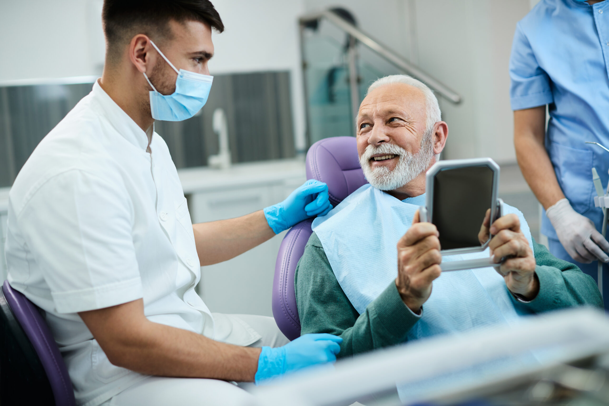 Satisfied senior man talks with dentist while looking his teeth in mirror at dentist's office