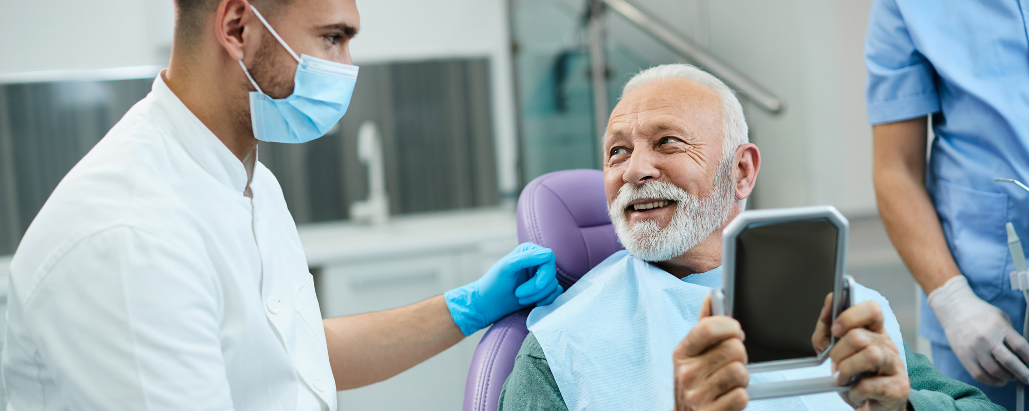 Satisfied senior man talks with dentist while looking his teeth in mirror at dentists office