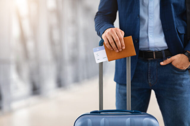 Ready For Trip. Unrecognizable Man Standing With Suitcase, Passport And Tickets At Airport Terminal, Male Traveller Waiting For Air Flight Near Boarding Gate, Cropped Image, Closeup Shot